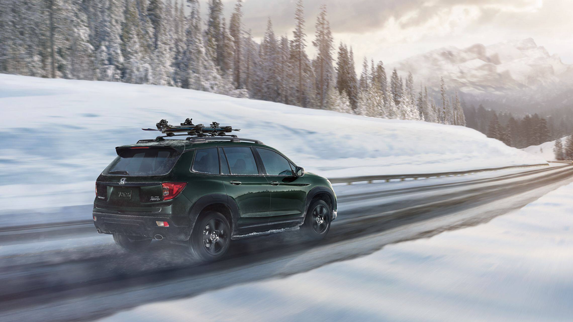Passenger-side rear 3/4 view of 2019 Honda Passport AWD Elite in Black Forest Pearl with accessory ski/snowboard attachment driving on a snowy mountain  road.